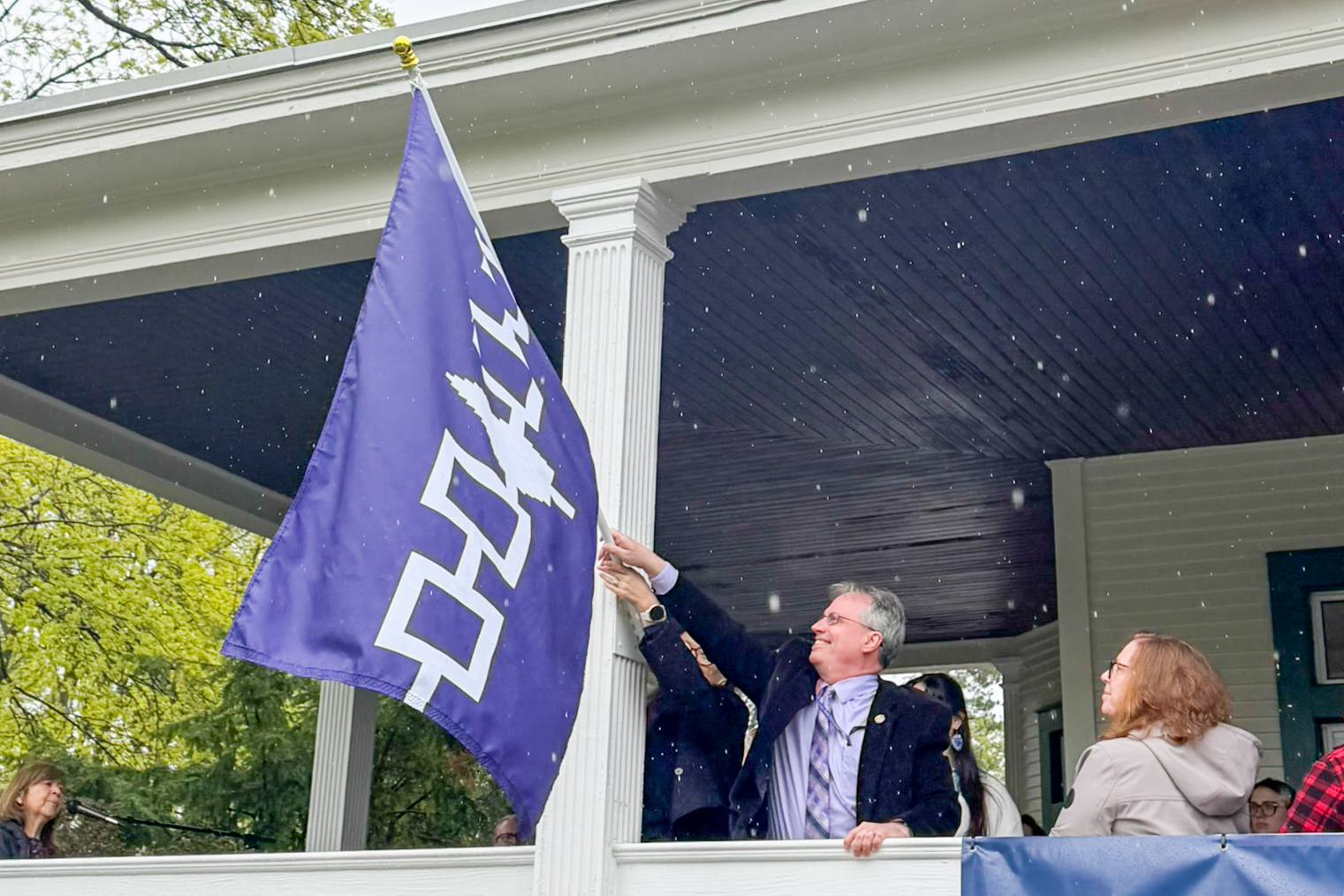 A man holds a flag pole that has a Haudenosaunee flag on it off of a porch with other people nearby