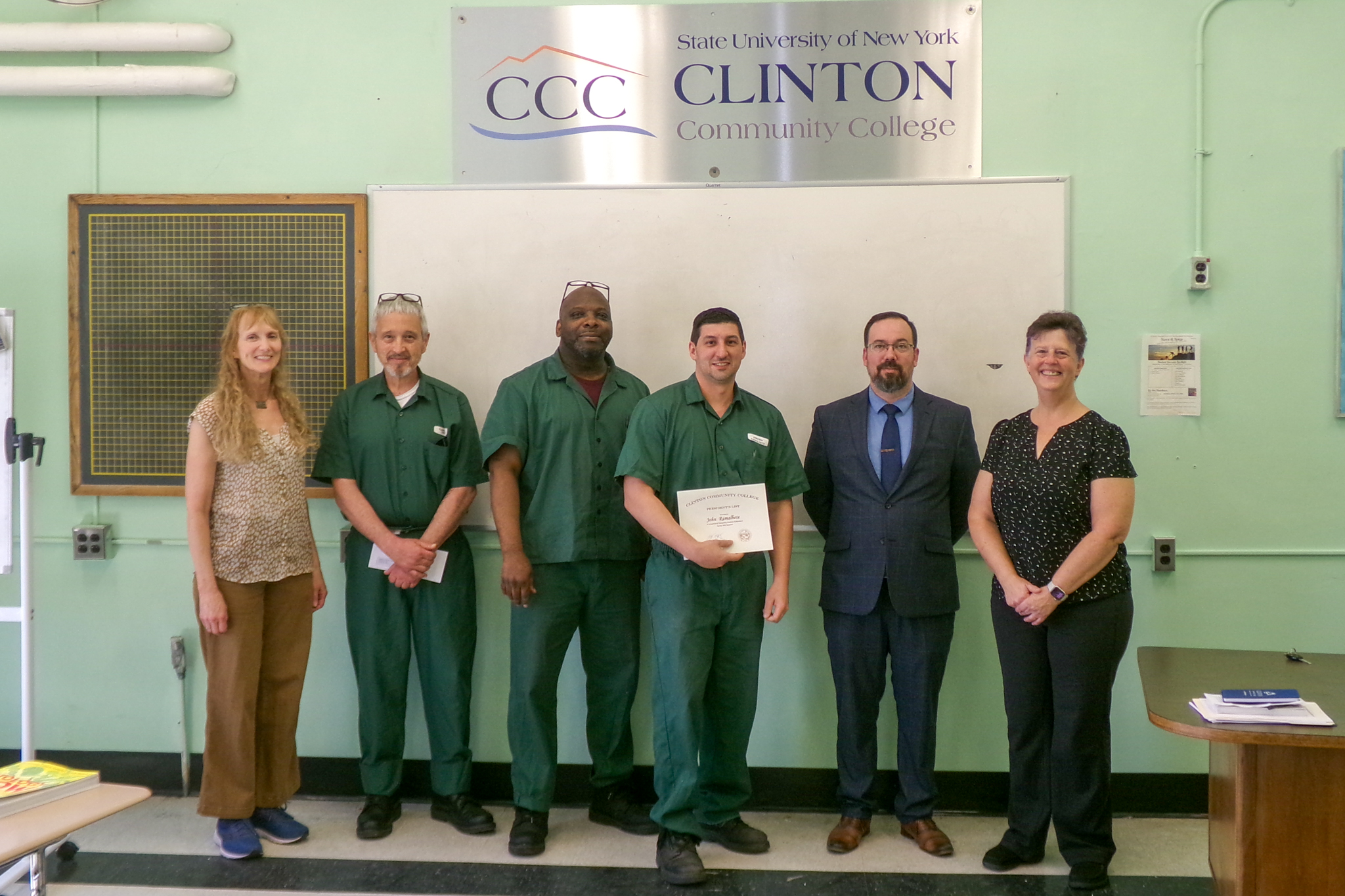 Six people standing side-by-side in a prison classroom with one person holding a certificate