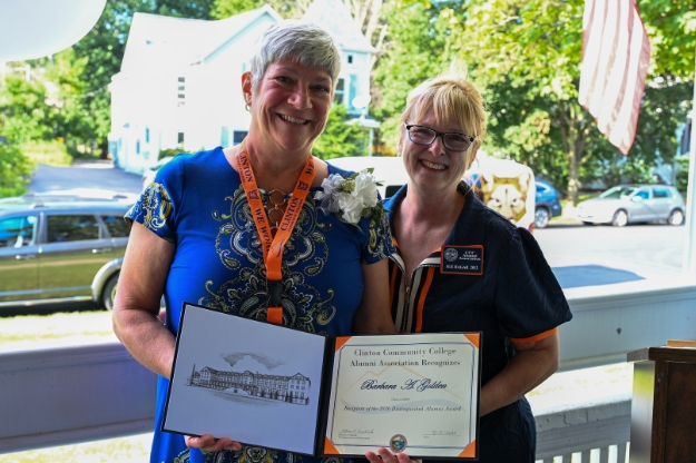 A woman holds an award while standing next to another woman on a porch