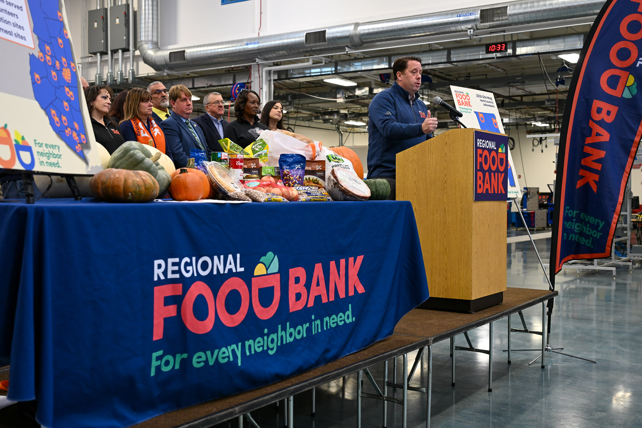 A man speaking at a podium next to a table with food on it and other people nearby