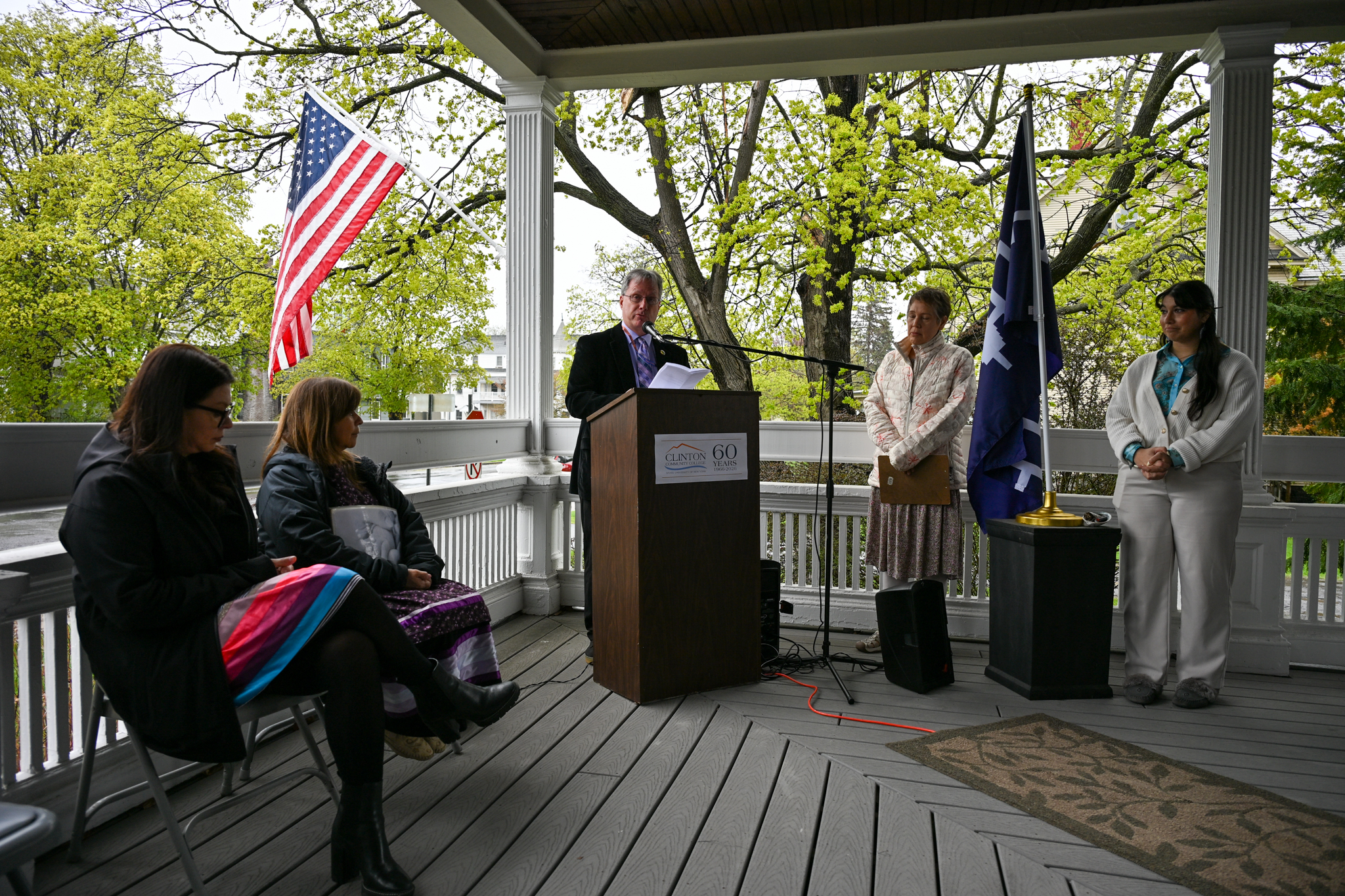 Several people on a porch with one behind a podium next to a Haudenosaunee flag
