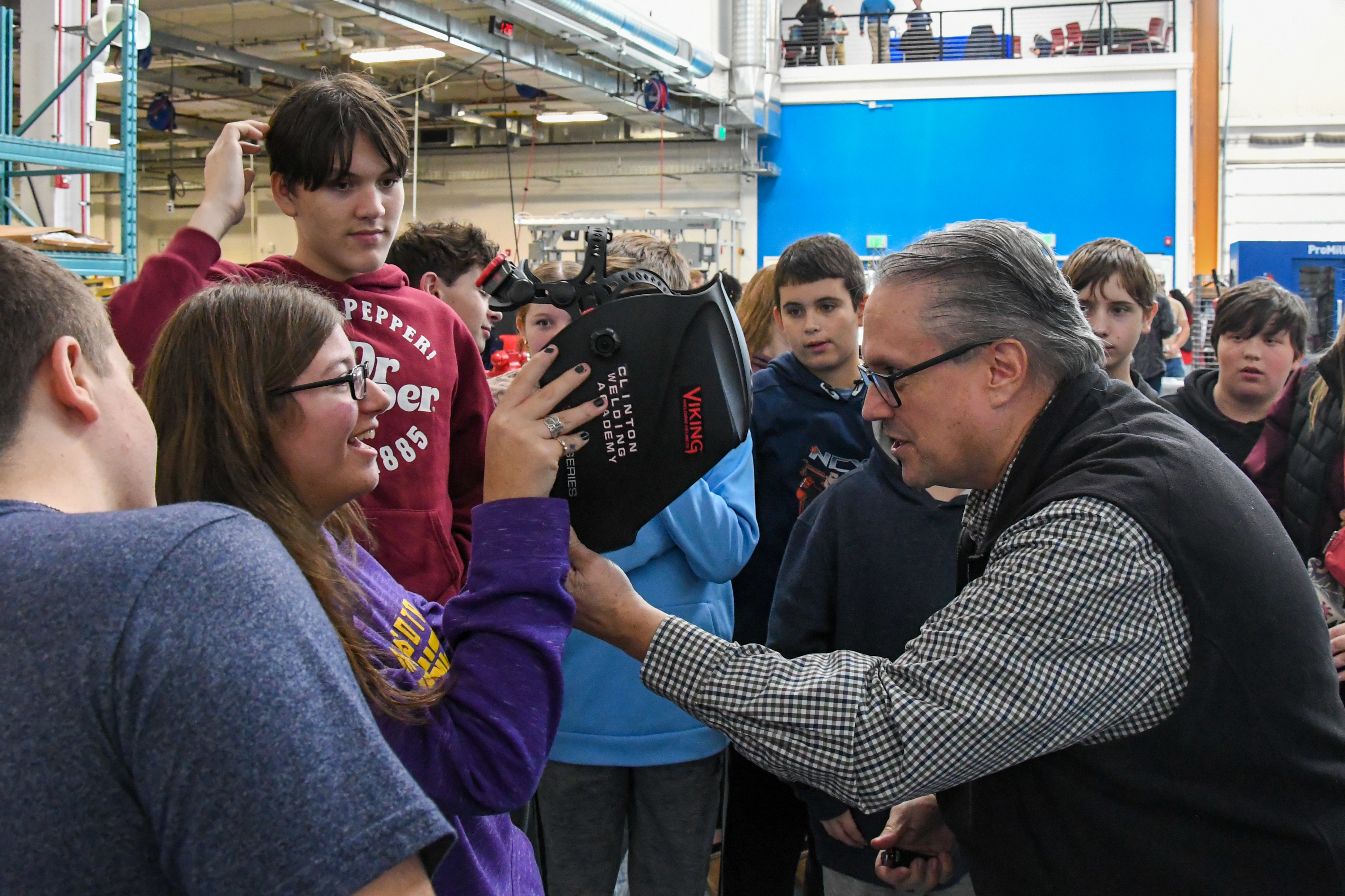 An instructor holds a welding helmet with a student about to put it on surrounded by a larger group of students