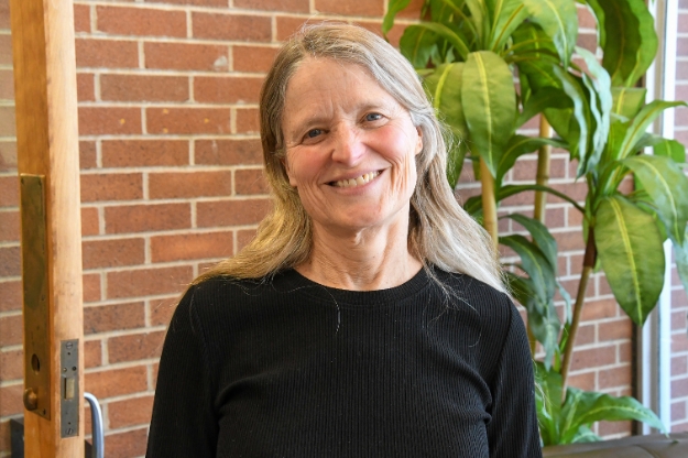 A woman smiling for a photo in front of a brick wall and green plant